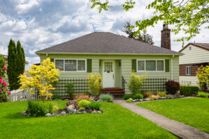 A single-story house with light green siding and a gray, shingled roof. There is a walkway going through the center of the grassy lawn, leading up to the three front steps. Part of another house is visible on the right.