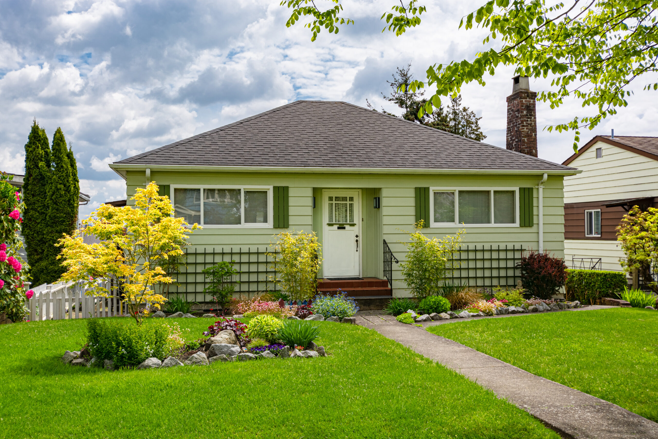 A single-story house with light green siding and a gray, shingled roof. There is a walkway going through the center of the grassy lawn, leading up to the three front steps. Part of another house is visible on the right.